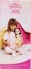 A young girl is sitting on the floor, holding a doll and a book. Anteprima dell’immagine prodotto