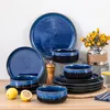 A collection of blue and black ceramic dishes, including plates and bowls, arranged on a wooden surface. A pepper grinder and a vase with yellow flowers are also visible. Voir les images du produit