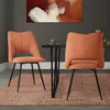 Two orange dining chairs are placed around a small dark wood table with a green cup on it. A painting hangs on the wall behind them.