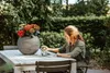 A woman sitting at a table outdoors with a vase of flowers. Náhľad obrázku produktu