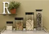 Four clear storage containers with black lids are arranged on a metal shelf against an olive green wall. The containers hold different food items: oats, pasta, peanuts, and dried herbs. A white letter 'R' and a potted plant are on a shelf above. Produktbild-Vorschau