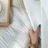 A hand is shown lifting a corner of a white quilted blanket. Náhľad obrázku produktu