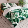 A bed with green and white banana leaf patterned bedding, set in a bedroom with wooden flooring and a plant in the background. Podgląd zdjęcia produktu