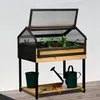 A wooden raised garden bed with a greenhouse lid, set up outdoors on a wooden deck. Accessories like a watering can and bucket are placed below. Produktbild-Vorschau