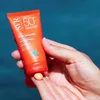 A hand holding an orange tube of sunscreen with a dollop of sunscreen on a finger, with a blue background. Náhled obrázku produktu