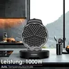 A waffle iron is shown open, displaying its heart-shaped waffle molds, on a kitchen counter. In the background, a modern kitchen backsplash and a window with a mountain view are visible. Text overlays provide product specifications. Náhled obrázku produktu
