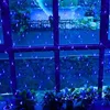 Blue net lights are draped across a window, with plants visible in the foreground and a reflection of trees and water in the background. Náhled obrázku produktu