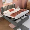 A bedroom scene with a grey upholstered bed frame that has its storage compartment open, revealing stored items. The bed is made with white linens and an orange throw blanket and pillow. There are decorative items on a side table and a chest of drawers in  Produktbild-Vorschau