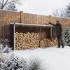 A man is placing firewood into a metal firewood storage unit next to a wooden fence in a snowy environment. Podgląd zdjęcia produktu