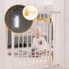 A baby is sitting on the floor near a white baby gate, playing with a toy car. An inset circle highlights a light and battery indicator on the gate. Náhľad obrázku produktu