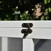 A close-up of a white wooden cage with a bronze latch, with green foliage and white flowers in the background. Náhled obrázku produktu