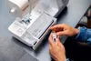 A person's hands are shown holding a bobbin and bobbin case near an open sewing machine. Produktbild-Vorschau