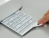 A hand is holding a white card with rows of stitch patterns printed on it, which is partially inserted under a white sewing machine. Produktbild-Vorschau