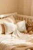 A close-up shot of a cream-colored bow-shaped pillow on a bed, with a teddy bear to the right. Podgląd zdjęcia produktu