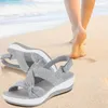 Paar graue Stoffsandalen mit weißen Sohlen an einem Sandstrand, im Hintergrund gehen Füße einer Person weg Anteprima dell’immagine prodotto