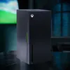 A black Xbox Series X replica mini-fridge is shown on a dark surface with a blurred background. Podgląd zdjęcia produktu