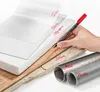A hand holding a black Staedtler Lumocolor permanent marker with a red cap, writing on various materials like film, glass, styrofoam, wood, stone, metal, and plastic. The materials are layered to demonstrate the marker's versatility. Produktbild-Vorschau