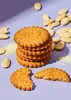 A stack of almond cookies with scattered almonds and almond crumbs on a light purple background. Produktbild-Vorschau