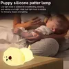 A person is feeding a baby while a puppy-shaped night light is on a table. Produktbild-Vorschau