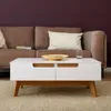A white coffee table with wooden accents is placed in front of a brown sofa. Produktbild-Vorschau