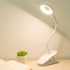 A white clip-on LED desk lamp with a flexible gooseneck is shown on a wooden surface next to a small potted plant and an open book. Produktbild-Vorschau