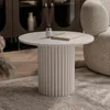 A round coffee table with a fluted base is placed on a rug in a living room setting, next to a textured armchair. A diffuser and a book are on the table. Voir les images du produit