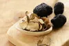 A wooden spoon filled with dried truffle slices sits on a wooden surface next to whole black truffles. Produktbild-Vorschau