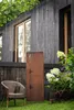 A decorative metal screen with a floral design stands in a garden next to a wicker chair and flowering plants. Produktbild-Vorschau