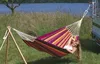 A woman relaxing in a colorful hammock supported by a wooden stand on one side and attached to a vehicle on the other, with a lake and trees in the background. Podgląd zdjęcia produktu