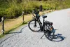 An electric bicycle parked on a gravel path next to a grassy embankment with a rope fence. Produktbild-Vorschau