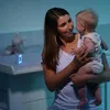A mother holds her baby in a dimly lit room, possibly a nursery. Voir les images du produit