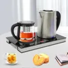 A tea set with a glass teapot and a stainless steel kettle on a warming plate, with a cup of tea and cookies in the foreground. Podgląd zdjęcia produktu