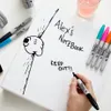 A hand is writing on a notebook with a black Sharpie marker. Other Sharpie markers and headphones are visible in the background. Voir les images du produit