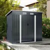 A dark gray metal shed with white trim is situated on a wooden deck in a garden setting, surrounded by greenery and houses. Produktbild-Vorschau