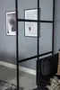 A modern living room interior with a black metal-framed glass partition, two framed art prints on the wall, and a black sofa with cushions. Produktbild-Vorschau
