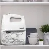 A white electric hand mixer is placed on a shelf next to some kitchenware and a plant. Produktbild-Vorschau