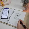 A person is using a smart pen to draw a panda in a notebook, with a smartphone displaying the same panda image next to it. Náhľad obrázku produktu