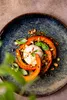 A close-up shot of a dish with roasted pumpkin slices arranged in a spiral around a ball of burrata cheese, garnished with basil leaves and chopped nuts. Náhľad obrázku produktu