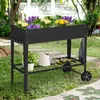 A raised garden bed filled with colorful flowers and plants, placed on a lawn in a garden setting. Gardening tools and a watering can are visible on a lower shelf. Voir les images du produit