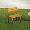 A wooden chair with metal legs sits on a green lawn in front of a white fence. Produktbild-Vorschau