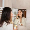 Frau in einem weißen Bademantel, die ein weißes und orangefarbenes Hautpflegeprodukt auf ihre Nase aufträgt, während sie in einen Spiegel schaut Anteprima dell’immagine prodotto