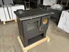 A black wood-burning stove with an oven and cooktop is displayed on a wooden pallet in what appears to be a warehouse or storage area. Other products and packaging are visible in the background. Produktbild-Vorschau