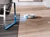 A white robot vacuum cleaner is cleaning a wooden floor next to a black leather chair with a silver frame. Voir les images du produit