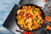 A close-up overhead shot of a pan filled with cooked vegetables, placed on a blue wooden surface with a black reflective surface and some floral elements. Produktbild-Vorschau