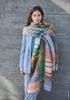 A woman stands in front of a gray wall wearing a blue jacket and a large, colorful striped scarf. Produktbild-Vorschau