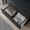 A drawer of a black dresser is open, showing folded clothes inside. A grey rug and wooden floor are visible. Náhled obrázku produktu