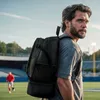 A man wearing a backpack stands on a sports field with another person in the background. Produktbild-Vorschau
