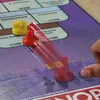 A hand is moving a red Monopoly game piece across a Monopoly board, passing the 'GO' space. Another yellow game piece is visible. Voir les images du produit
