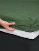 A hand is shown tucking a green fitted sheet onto a mattress. Produktbild-Vorschau
