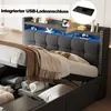 A close-up view of a grey upholstered bed with storage underneath, featuring LED lighting and a shelf with a tablet and charging accessories. Produktbild-Vorschau